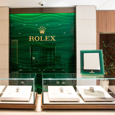 The Rolex Showroom at Michael Spiers in Exeter. The Rolex logo is visible on the wall with 3 Rolex watches visible in the foreground in glass cases.